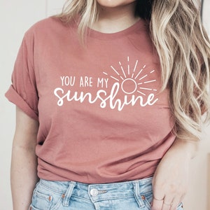 May include: A dusty rose t-shirt with rolled-up sleeves. The shirt features the white text "YOU ARE MY sunshine" with a sun graphic. The model is wearing blue jeans.
