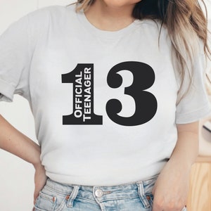 May include: A white t-shirt with the text "13 OFFICIAL TEENAGER" printed in black. The number 13 is large and bold, with the words "OFFICIAL TEENAGER" stacked vertically to the left of the number.