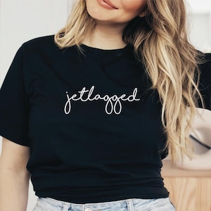 以下が含まれることがあります： 白い筆記体で「jetlagged」という文字が入った黒いクルーネックTシャツ。明るいブルーデニムのジーンズと合わせています。モデルはシャツとジーンズを着ています。