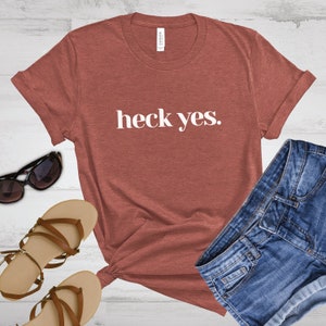 May include: A rust-colored t-shirt with the phrase "heck yes." in white block letters. The shirt is styled with denim shorts, sunglasses, and sandals. The t-shirt is made of a soft material.
