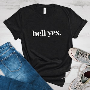 May include: Black t-shirt with the phrase "hell yes." in white, paired with blue jeans and gray canvas sneakers. The shirt is made of a soft material and has rolled-up sleeves.
