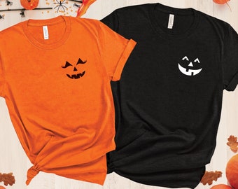 Puede incluir: Dos camisetas, una naranja y otra negra, cada una con un diseño de cara de calabaza. La camiseta naranja tiene una cara negra, mientras que la camiseta negra tiene una cara blanca. Las camisetas se muestran con decoraciones de Halloween.