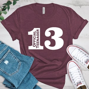 May include: A maroon t-shirt with the text "OFFICIAL TEENAGER 13" in white. The shirt is paired with blue jeans and white sneakers. The t-shirt is a casual wear item.