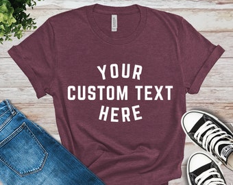 May include: A maroon t-shirt with white text that reads "YOUR CUSTOM TEXT HERE". The shirt is laid flat on a wooden surface with a pair of black sneakers and blue jeans.