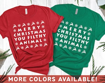 Könnte beinhalten: Rotes und grünes T-Shirt mit dem weißen Text "MERRY CHRISTMAS YOU FILTHY ANIMAL". Das Design umfasst Rentiere und Schneeflocken. Der Schriftzug "MORE COLORS AVAILABLE!" befindet sich unten.