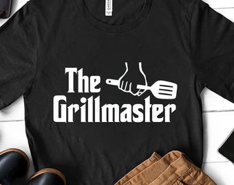 May include: Black t-shirt with the text "The Grillmaster" in white, featuring a graphic of a hand holding a spatula. The shirt is displayed with brown pants, black shoes, and a leather bag on a white wooden surface.