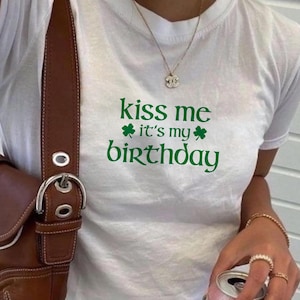 May include: White t-shirt with green text that reads "kiss me it's my birthday" with shamrock symbols.