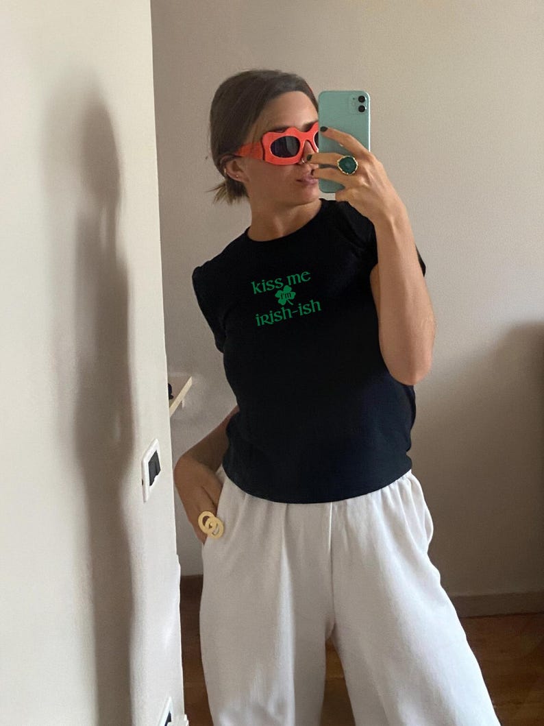 May include: A black t-shirt with the text "kiss me I'm irish-ish" and a green shamrock graphic. The person is wearing red heart-shaped sunglasses and a gold ring.