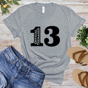 May include: A heather gray t-shirt with the text "13 OFFICIAL TEENAGER" in black. The shirt is displayed with blue denim shorts and brown sandals on a wooden surface. A small green plant is also visible.