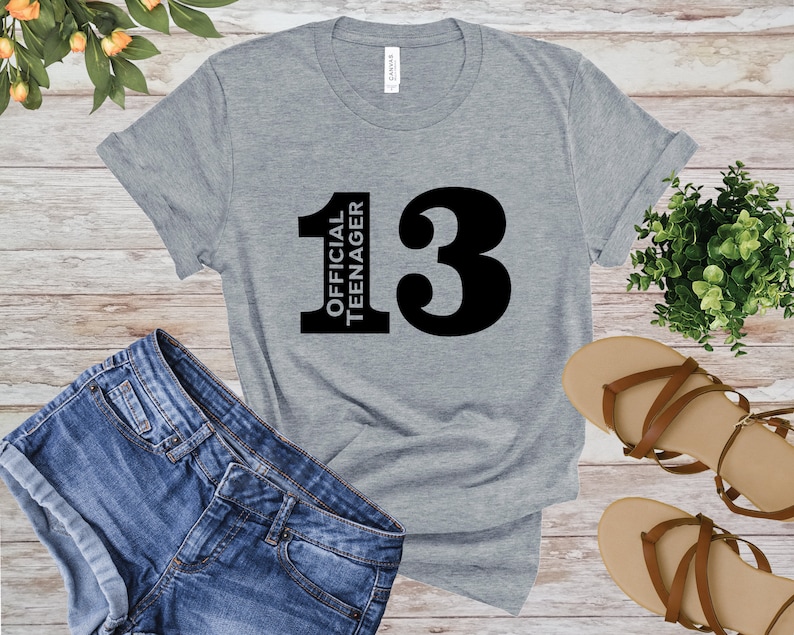 May include: A heather gray t-shirt with the text "13 OFFICIAL TEENAGER" in black. The shirt is displayed with blue denim shorts and brown sandals on a wooden surface. A small green plant is also visible.