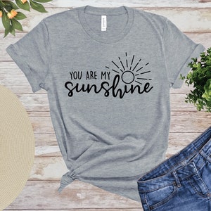 May include: A grey t-shirt featuring the words "YOU ARE MY sunshine" in black script, with a sun graphic. The shirt is styled with denim shorts and a straw hat, on a wooden surface. The shirt is made of a soft material.