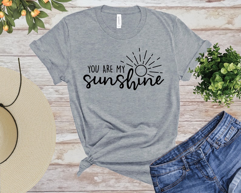 May include: A grey t-shirt featuring the words "YOU ARE MY sunshine" in black script, with a sun graphic. The shirt is styled with denim shorts and a straw hat, on a wooden surface. The shirt is made of a soft material.