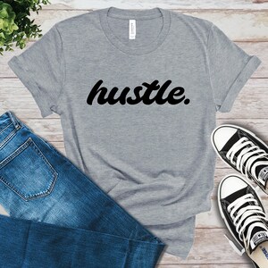 May include: A gray t-shirt with the word "hustle" in black script. The shirt is paired with blue jeans and black and white sneakers, all arranged on a light wood-grain surface. The shirt's sleeves are rolled up.