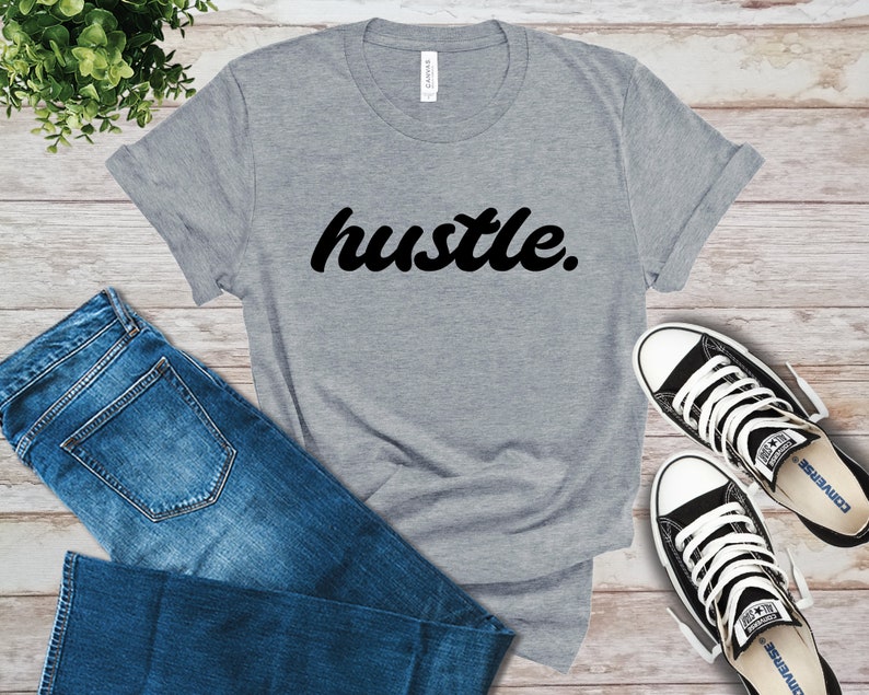 May include: A gray t-shirt with the word "hustle" in black script. The shirt is paired with blue jeans and black and white sneakers, all arranged on a light wood-grain surface. The shirt's sleeves are rolled up.