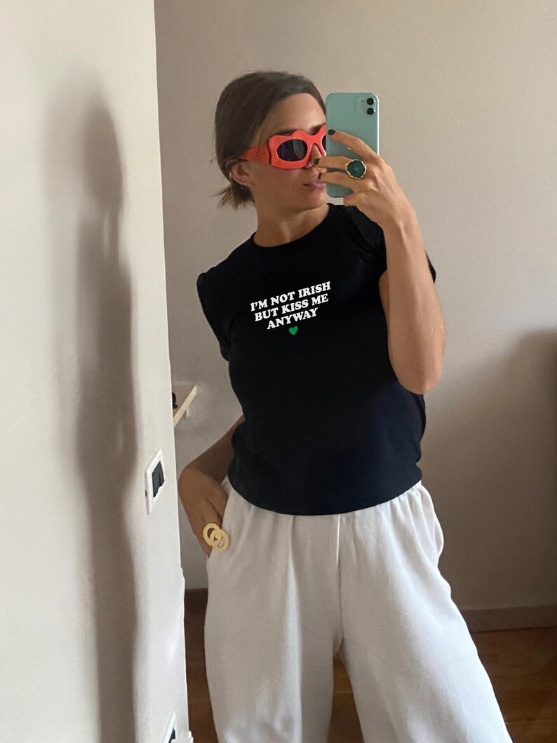 May include: A black t-shirt with the text "I'm not Irish but kiss me anyway" in white with a green heart. The person is wearing red sunglasses and a gold ring.