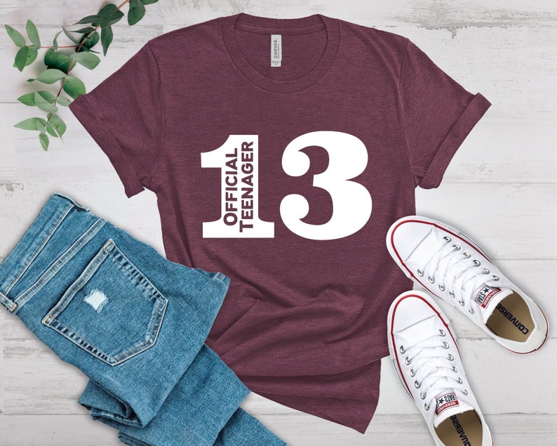 May include: A maroon t-shirt with the text "OFFICIAL TEENAGER 13" in white. The shirt is paired with blue jeans and white sneakers. The t-shirt is a casual wear item.