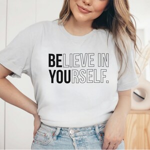 May include: A light gray t-shirt with the text "BELIEVE IN YOURSELF" in black and outlined letters. The shirt is made of a soft material and has short sleeves. The shirt is a casual wear item.