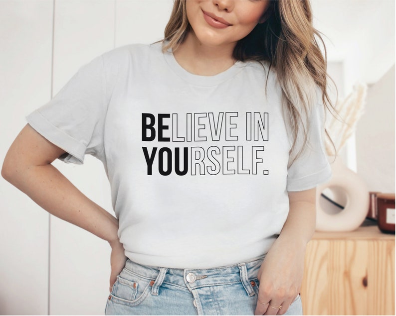 May include: A light gray t-shirt with the text "BELIEVE IN YOURSELF" in black and outlined letters. The shirt is made of a soft material and has short sleeves. The shirt is a casual wear item.