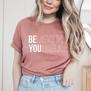 May include: A dusty rose t-shirt with the text "BELIEVE IN YOURSELF" in white block letters. The shirt has short sleeves and is made of a soft material. The model is wearing blue jeans.