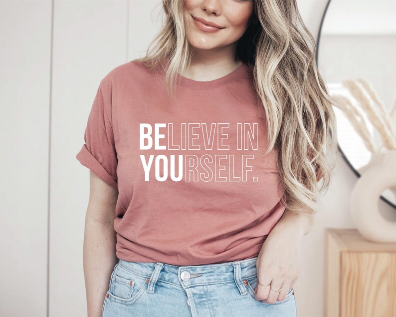 May include: A dusty rose t-shirt with the text "BELIEVE IN YOURSELF" in white block letters. The shirt has short sleeves and is made of a soft material. The model is wearing blue jeans.