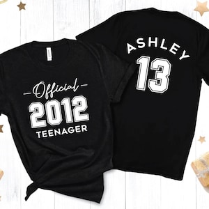 Pode incluir: Duas camisetas pretas com texto branco. Uma camiseta diz "Oficial 2012 Adolescente" e a outra diz "Ashley 13".