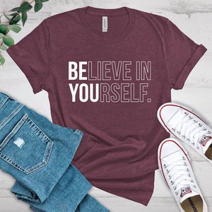 May include: Maroon t-shirt with the text "BELIEVE IN YOURSELF" in white. The shirt is paired with blue denim jeans and white canvas sneakers with red accents. The items are arranged on a light-colored wooden surface.