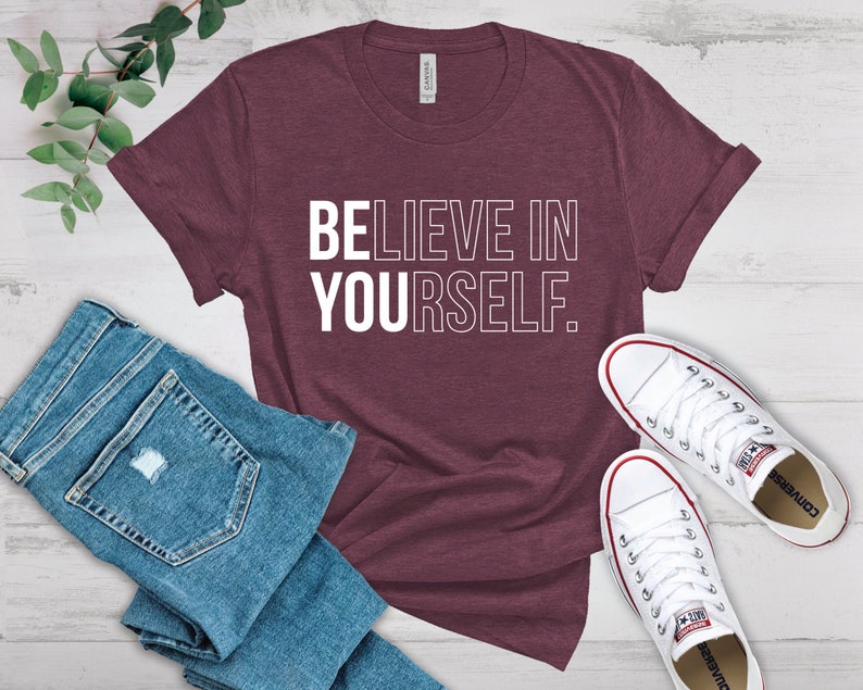 May include: Maroon t-shirt with the text "BELIEVE IN YOURSELF" in white. The shirt is paired with blue denim jeans and white canvas sneakers with red accents. The items are arranged on a light-colored wooden surface.