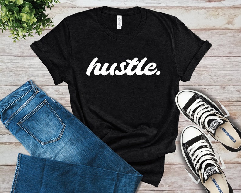 May include: Black t-shirt with the word "hustle" in white, cursive font. The shirt is paired with blue jeans and black and white Converse sneakers. The items are arranged on a light wood-grain surface.