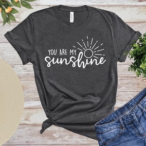 May include: Dark grey t-shirt featuring the white text "YOU ARE MY sunshine" and a sun graphic. The shirt is knotted at the hem and is shown with denim shorts and a straw hat. The shirt is made of a soft material.