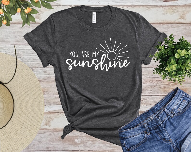 May include: Dark grey t-shirt featuring the white text "YOU ARE MY sunshine" and a sun graphic. The shirt is knotted at the hem and is shown with denim shorts and a straw hat. The shirt is made of a soft material.