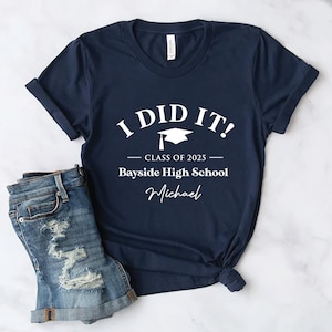 以下が含まれることがあります： 「I DID IT！」の文字と卒業帽子のグラフィックが入ったネイビーブルーのTシャツ。「CLASS OF 2025」、「Bayside High School」、そして名前の「Michael」が下に書かれています。Tシャツはデニムのショートパンツと組み合わせています。