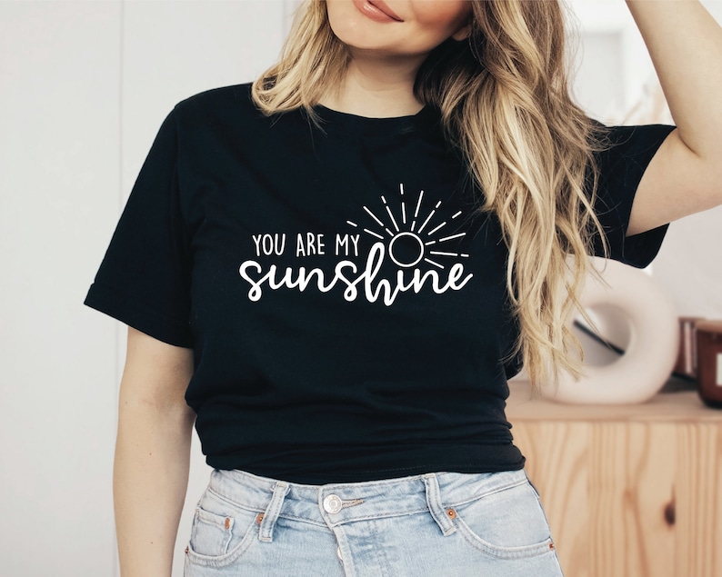 May include: Black t-shirt featuring the white text "YOU ARE MY sunshine" and a sun design. The t-shirt has a crew neck and is made of a soft fabric. The shirt is worn by a person.