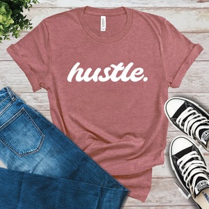 May include: A dusty rose t-shirt with the word "hustle" in white script. The shirt is paired with blue jeans and black and white sneakers. The items are arranged on a light wood-grain surface.