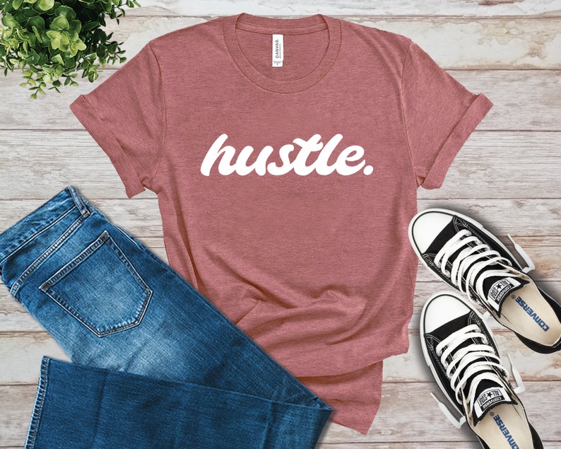 May include: A dusty rose t-shirt with the word "hustle" in white script. The shirt is paired with blue jeans and black and white sneakers. The items are arranged on a light wood-grain surface.