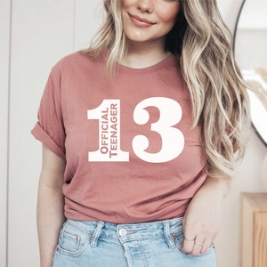 May include: A dusty rose-colored t-shirt with the text "OFFICIAL TEENAGER" stacked vertically next to the number "13" in white. The shirt is a casual style, with rolled-up sleeves.