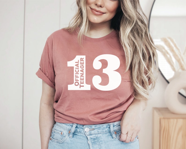 May include: A dusty rose-colored t-shirt with the text "OFFICIAL TEENAGER" stacked vertically next to the number "13" in white. The shirt is a casual style, with rolled-up sleeves.