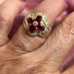 May include: A gold ring with a floral design. The ring features four deep red gemstones at the center, surrounded by smaller white gemstones and baguette-cut stones. The ring is set on a gold band.