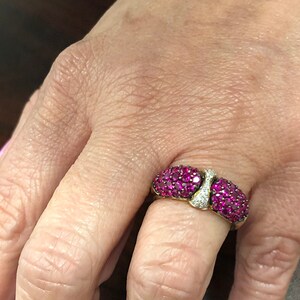 May include: A gold ring with a band of small pink gemstones and a center design of larger pink gemstones. The ring is worn on a person's finger.