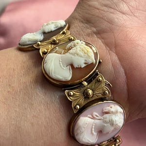 May include: A gold-toned cameo bracelet featuring oval cameos with white relief portraits set in ornate gold frames. The bracelet is connected by decorative gold links and a green stone accent.
