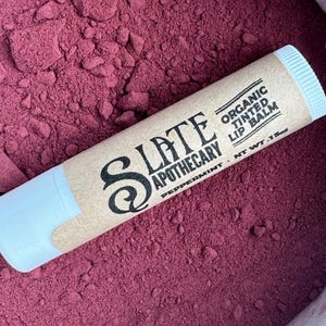 May include: A brown tube of Slate Apothecary Organic Tinted Lip Balm in Peppermint. The tube is lying on a bed of deep red powder.
