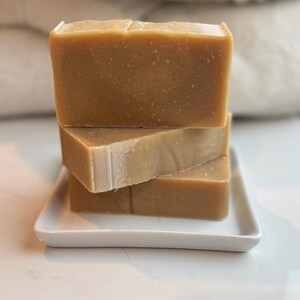 May include: Three bars of handmade soap stacked on a white ceramic dish. The soap is a light brown color and has a natural, rustic look.