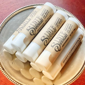 May include: A collection of organic lip balms in a silver dish. The lip balms are in white tubes with brown paper labels. The labels read "Slate Apothecary Organic Lip Balm Peppermint NT WT .15oz". The dish sits on a red surface.