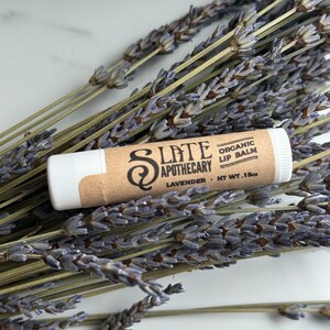 May include: A white tube of organic lip balm with a brown paper label that says "Slate Apothecary" and "Lavender - Net Wt. .15oz". The tube is sitting on a bunch of dried lavender flowers.