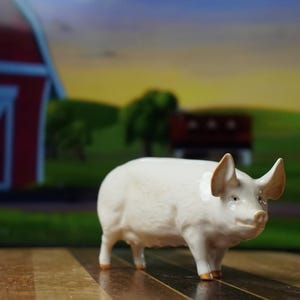 May include: A ceramic pig figurine with a white body, tan ears, and hooves. The pig is standing on a wooden surface, with a painted farm scene in the background, including a red barn and green fields.