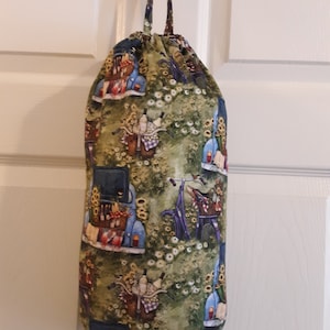 May include: A green fabric bag with a drawstring closure. The bag is decorated with a repeating pattern of blue cars, bicycles, and baskets filled with fruit and flowers. The bag is hanging on a white door.