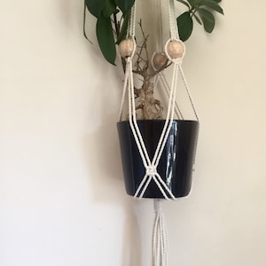 Pode incluir: Um suporte de planta de macramê branco com contas de madeira e um vaso preto com uma planta dentro.