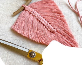 May include: A pink macrame feather wall hanging with a wooden branch at the top. The feather is made with pink yarn and has a fringe at the bottom. The feather is hanging on a light brown fabric background.