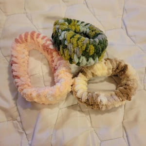 Puede incluir: Tres scrunchies para el pelo de crochet de colores. Uno es rosa y blanco, otro es marrón y blanco, y el último es verde, blanco y gris.