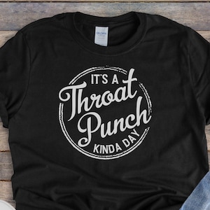 Może przedstawiać: Czarny T-shirt z białym kołem graficznym, na którym widnieje napis "It's a Throat Punch Kinda Day". Koszulka leży na drewnianej powierzchni z parą białych butów sportowych i niebieskimi dżinsami.