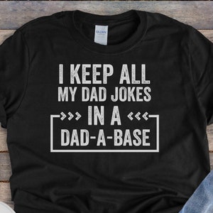 Op de afbeelding: Zwart T-shirt met witte tekst die "I keep all my dad jokes in a dad-a-base" luidt. Het shirt ligt plat op een houten oppervlak met een paar witte sneakers en een blauwe jeans.